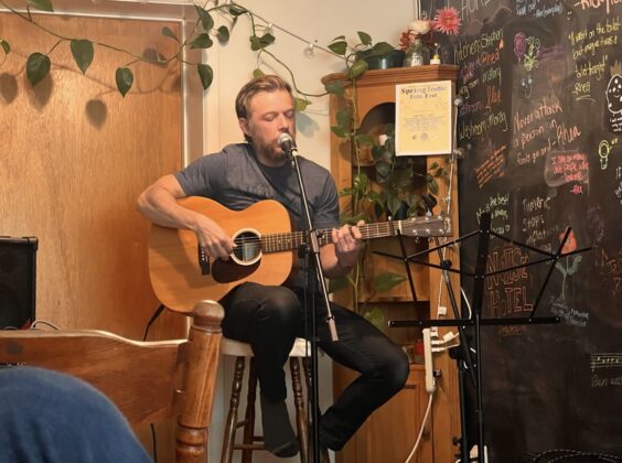 A person sitting on a stool behind a mic stand plays guitar and sings in a living room.
